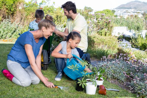 A family of four engaging in gardening activities in a well-maintained garden, with lush green grass, flowering plants, and shrubbery visible in the background. The foreground features a woman kneeling on the lawn using a small gardening fork near a young plant; nearby are gardening tools, a potted plant, and a watering can. A young girl, sitting on the grass, helps with planting, holding a small plant, while an older man and a young boy stand behind them, observing and smiling. The garden appears to be a private outdoor space with vibrant flower beds, leafy shrubs, and a backdrop of distant trees and a partly cloudy sky, illuminated by natural daytime light, reflecting a peaceful, family-friendly outdoor setting. This scene exemplifies typical garden maintenance and family outdoor activities supported by local gardening services in Tufnell Park.