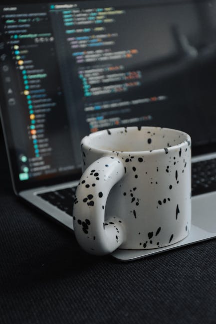 A close-up of a white ceramic mug with black splatter patterns on a black surface, in front of an open laptop displaying lines of code in a dark-themed text editor. The scene suggests a workspace setu