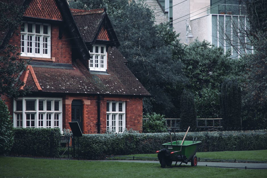 A traditional red-brick house with a steep, tiled roof and white-framed windows is set within a well-maintained garden. The garden features neatly trimmed hedges and lush green grass, with a small law